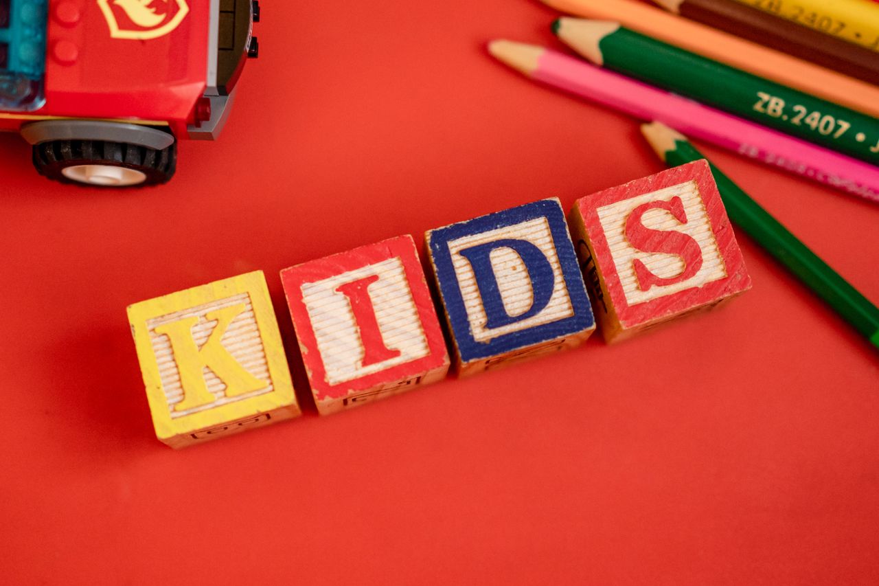 who-we-are Creative layout of wooden blocks spelling 'KIDS' with pencils and toy on red background.